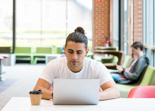 person sitting indoors at table with laptop and takeaway coffee