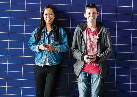 A young male and female stand in front of tiled blue wall and smile to camera; each is holding a smartphone in both hands. A young male and female stand in front of tiled blue wall and smile to camera; each is holding a smartphone in both hands.