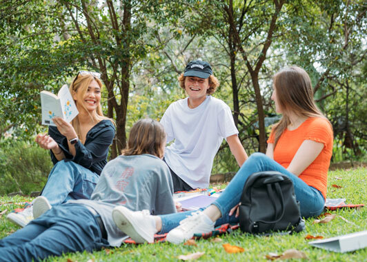 Four university students wearing jeans and t-shirts sit on the grass, talking and smiling, with trees behind them.