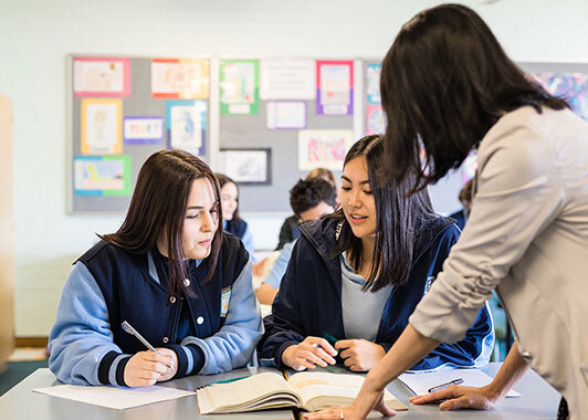 Woman facing away from camera leans towards two school students at their desks; books are open and both students are holding a pen