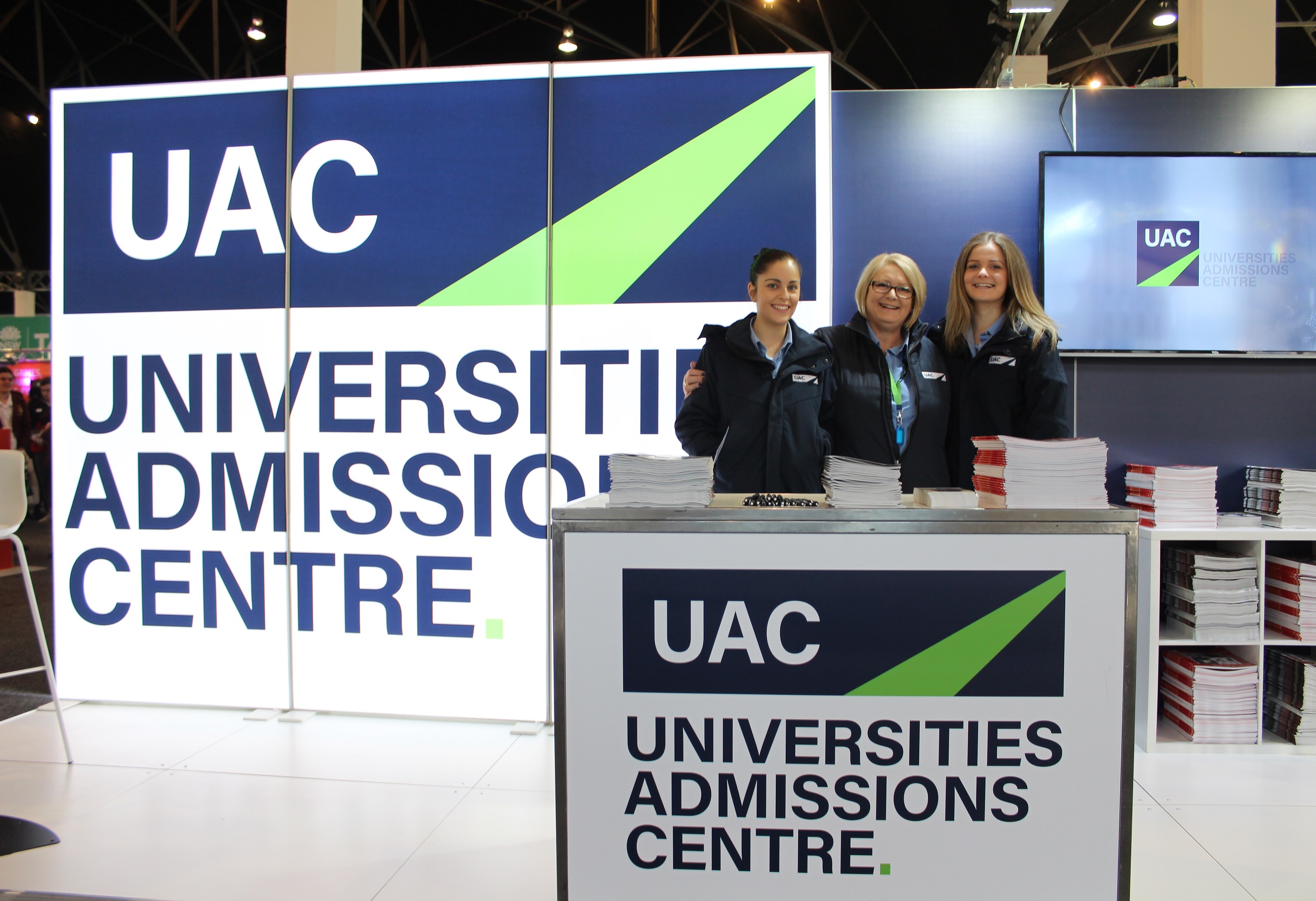 Three female UAC staff members stand smiling to camera at UAC expo stall; stacks of publications fill the stall shelves. Three female UAC staff members stand smiling to camera at UAC expo stall; stacks of publications fill the stall shelves.