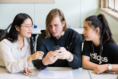 Three students in a classroom looking at a mobile phone