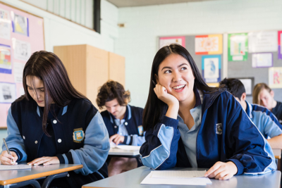 Classroom scene in which students, sitting at desks in rows, are busy working; the female student closest to camera is looking diagonally upwards smiling Classroom scene in which students, sitting at desks in rows, are busy working; the female student closest to camera is looking diagonally upwards smiling