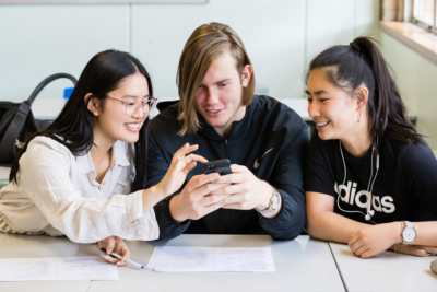 Three young people, a guy and two girls, huddle over a mobile phone smiling Three young people, a guy and two girls, huddle over a mobile phone smiling