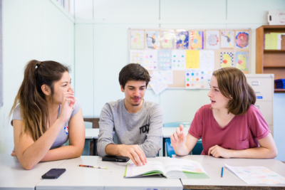 Three young people sitting at a table with pens, a smartphone, an open book, chatting Three young people sitting at a table with pens, a smartphone, an open book, chatting