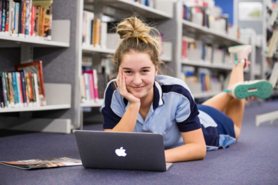 Female school student in uniform with laptop lying on floor of library and smiling to camera Female school student in uniform with laptop lying on floor of library and smiling to camera