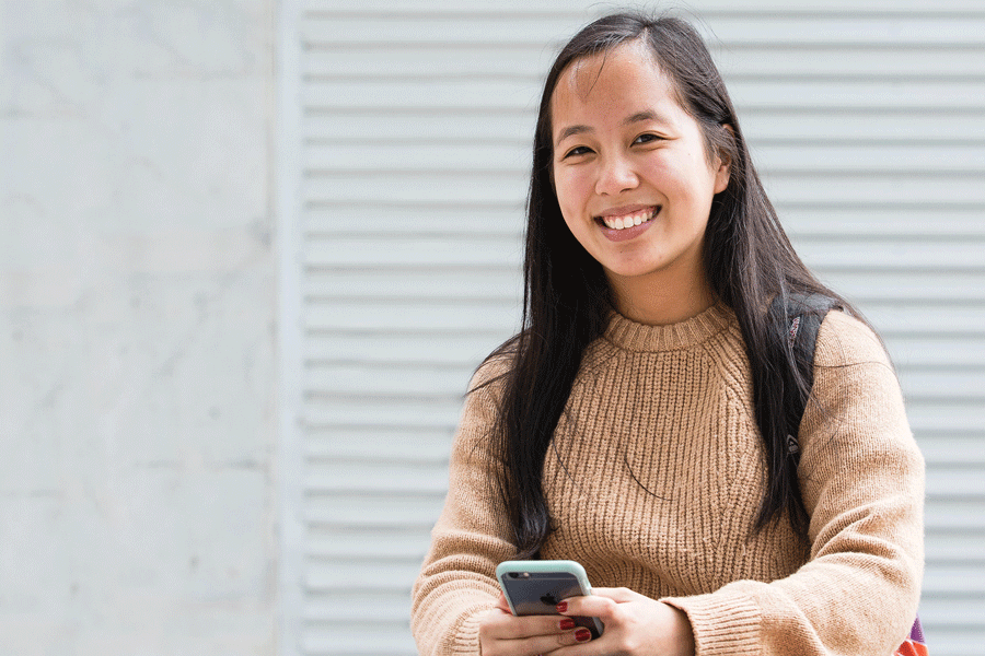Girl in tan coloured jumper holds mobile phone and smiles to camera in front of white wall Girl in tan coloured jumper holds mobile phone and smiles to camera in front of white wall