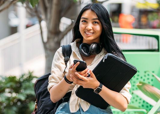 Female student smiles at camera while holding phone and laptop