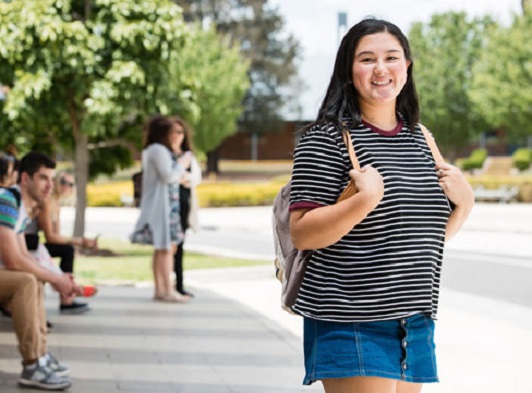 Girl standing outdoors facing camera holding backpack, smiling and wearing striped t-shirt Girl standing outdoors facing camera holding backpack, smiling and wearing striped t-shirt