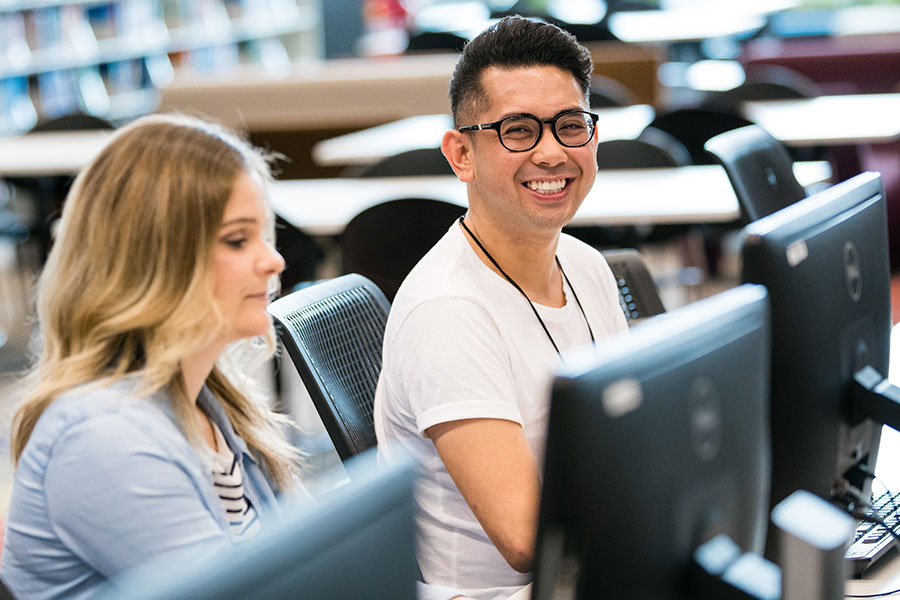 Guy with glasses next to girl smiles to camera sitting at one of a row of desktop computers Guy with glasses next to girl smiles to camera sitting at one of a row of desktop computers
