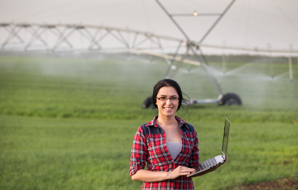 Young woman with laptop outside in check shirt and glasses standing in front of a large irrigation system in a field Young woman with laptop outside in check shirt and glasses standing in front of a large irrigation system in a field