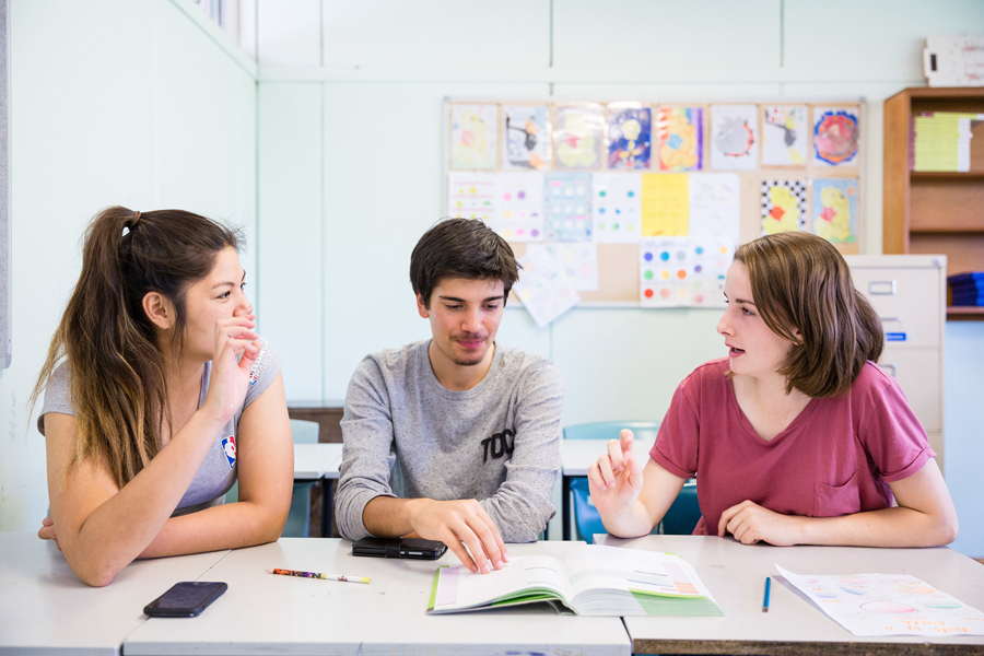 Three young people sitting at a table with pens, a smartphone, and an open book, are chatting and gesturing to each other Three young people sitting at a table with pens, a smartphone, and an open book, are chatting and gesturing to each other