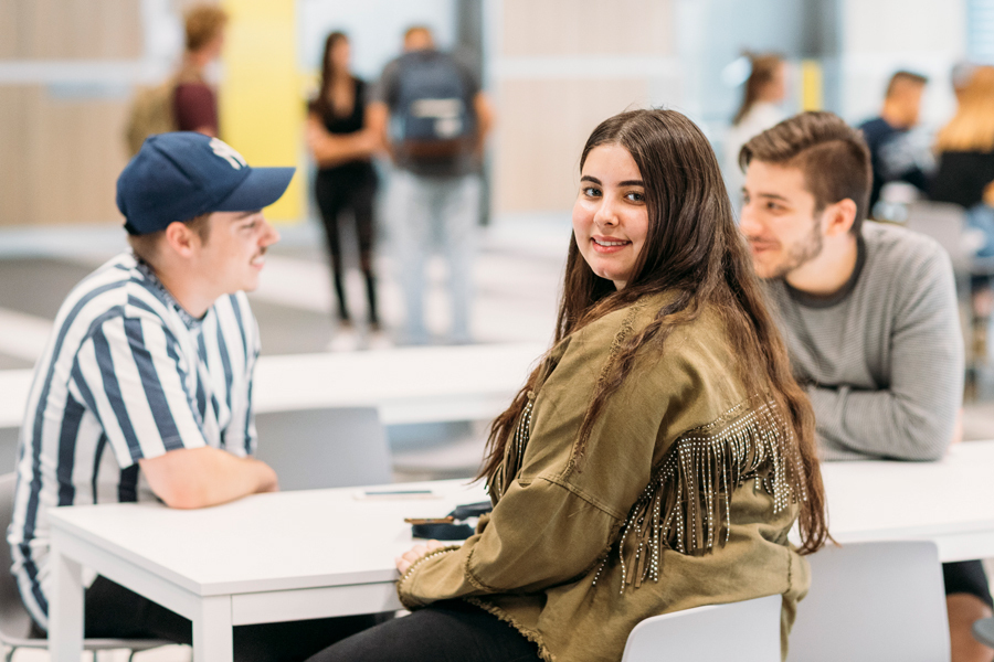 A young woman sits at a table with two male friends and smiles to camera