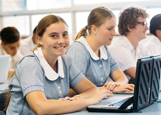 Two female high school students in a classroom using laptops. One student looks at the camera.
