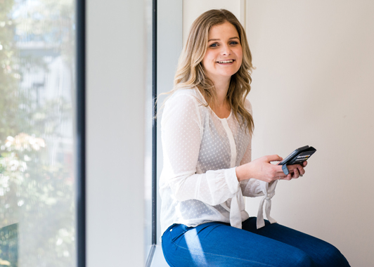 Young woman with long hair sitting near a window holding a smartphone and smiling to camera